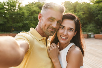 Portrait of happy middle-aged couple taking selfie photo and hugging while walking in summer park