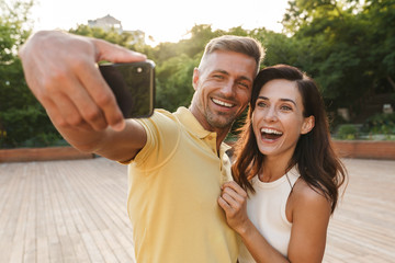 Portrait of excited adult couple taking selfie photo on cellphone and laughing while walking in summer park