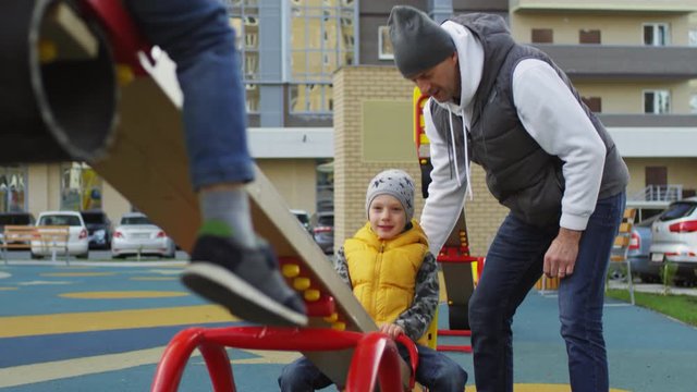 Tilting shot of 5-year-old Caucasian boy in warm vest, jeans and hat playing on seesaw with friend in communal playground of residential apartment block, and caring 40-something dad minding him