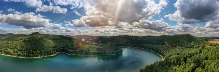 Camera flies over the Hennesee near Meschede and takes a wide view picture. You can see a ship, hills and forests, sunbeams and lens flares. Gorgeous nature in the Sauerland. - obrazy, fototapety, plakaty