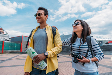 Woman in sunglasses holding digital camera near bi-racial friend with map and backpack