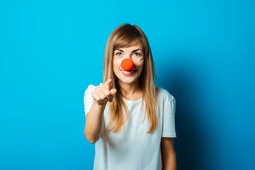 Beautiful young woman in a white T-shirt and a red clown nose smiles and points a finger on a blue...