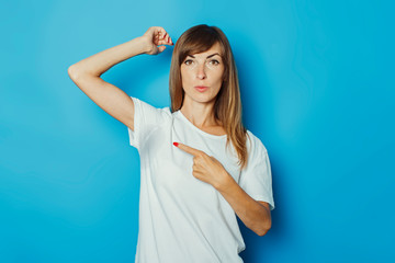 Young girl in a white T-shirt shows a finger on wet armpits from sweat on a blue background. Concept of excessive sweating, heat, deodorant - obrazy, fototapety, plakaty