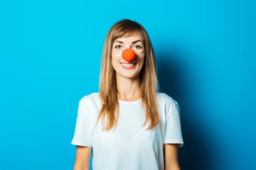 Beautiful young woman in a white T-shirt and red nose of a clown on a blue background. Concept party, costume, red nose day