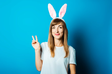 Young woman in bunny ears makes a hand gesture on a blue background. Easter concept