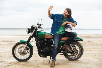 Fototapeta premium Handsome optimistic cheery man biker on his bike outdoors on a beach waving to friends.