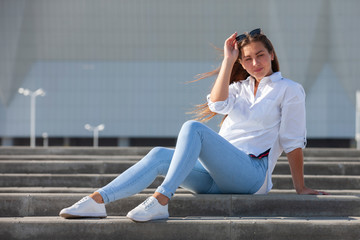 Fototapeta na wymiar Young American woman in a white T-shirt in jeans in white trendy sneakers relaxes sitting on the steps. Cute european girl model enjoys the rest. Spring style women's clothing.