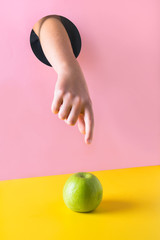 Childs hand through hole in pink paper reaches for green apple on yellow.Food concept.