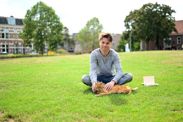 Naklejka na ściany i meble Spontaneous and happy young man sitting in a suburban park with a book and is caressing his cat