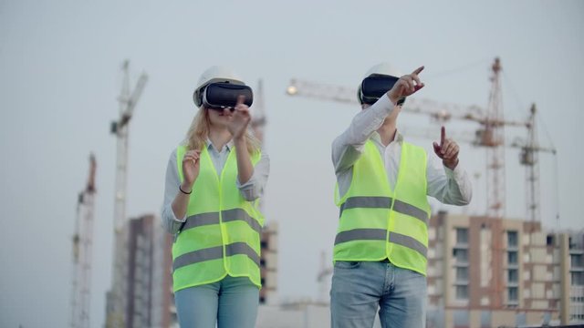 Two inspectors of the future on the construction site use virtual reality glasses on the background of buildings and cranes.