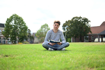 Happy and smiling young man reading a book in a small green park in the Netherlands