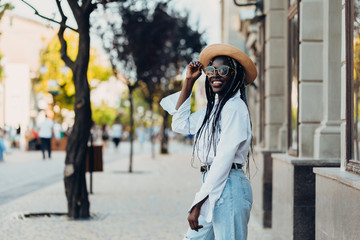 Portrait of a smiling young African American girl with pigtails in a fashionable hat and stylish sunglasses walking along the street on a sunny day