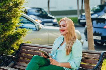 Fototapeta na wymiar Portrait happy smile beautiful elegant woman blue blouse sitting on a bench with a tablet in her hands. street portrait