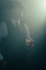 Retro businessman with cap holds glass of whiskey in smoky room.