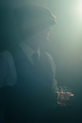 Retro businessman with cap holds glass of whiskey in smoky room.