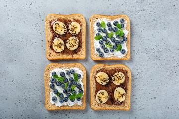 Delicious open sandwiches with goat cheese and blueberries and chocolate and banana on gray table. Breakfast background. Copy space, top view