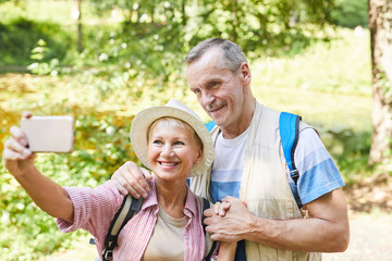 Happy mature woman standing and making selfie portrait with her husband on mobile phone while they standing in the park