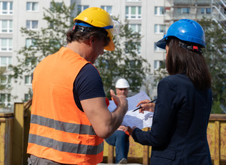 Construction manager wearing safety jacket and helmet checking projects discussing with a female engineer. In background, another worker