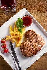 Grilled pork steak with fries, sauce and vegetables on white plate. Glass of red wine