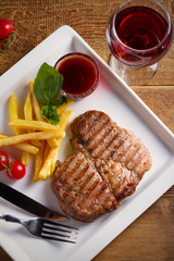 Grilled pork steak with fries, sauce and vegetables on white plate. Glass of red wine