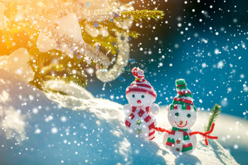 Two small funny toys baby snowman in knitted hats and scarves in deep snow outdoors near pine tree branch. Happy New Year and Merry Christmas greeting card.