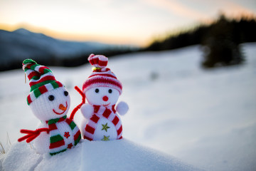 Two small funny toys baby snowman in knitted hats and scarves in deep snow outdoors on blurred mountains landscape background. Happy New Year and Merry Christmas greeting card theme.