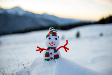 Small funny toy baby snowman in knitted hat and scarf in deep snow outdoor on blurred snow covered mountains landscape background. Happy New Year and Merry Christmas greeting card theme.