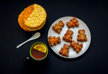 Homemade biscuits, bear shape candies with honey, sesame and sunflower seeds on a plate and cup of tea with lemon slice on a black background. Sweet breakfast or snack.