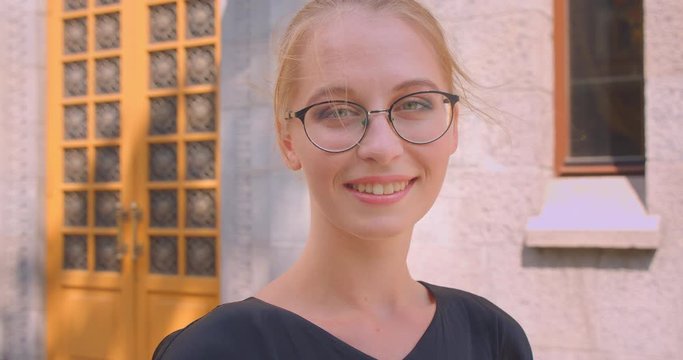 Closeup portrait of young attractive caucasian female in glasses looking at camera smiling cheerfully outdoors on street in city
