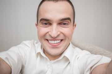 Fototapeta premium Handsome man in white shirt smiling for selfie, sitting at home on the couch, front view, portrait, close up, toned