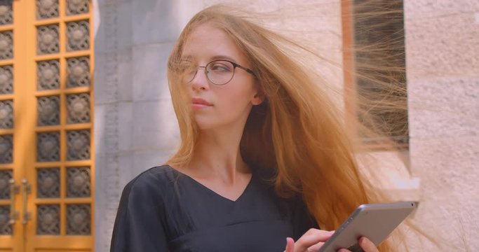Closeup portrait of young attractive caucasian female with long hair in glasses using tablet looking at camera outdoors on street