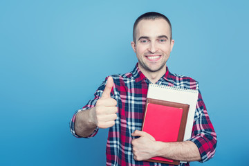 Happy student with books, man shows big thumb up, gesture super! Portrait, blue background, copy space, slogan, toned