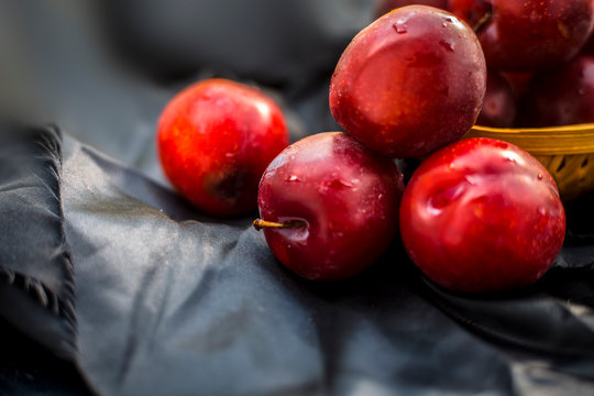 Raw organic red ripe plums in a brown-colored basket on black fabric on the wooden surface.