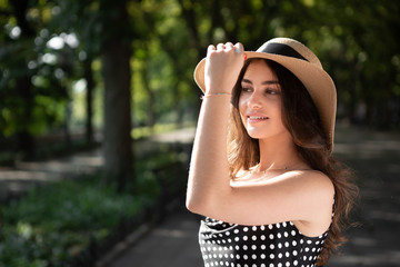 Close-up porttrait of a beautiful young brunette woman dressed in a black dress and a hat with wide flaps takes a walk in a park during warm summer day enjoying sunlight.