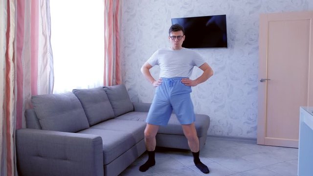 Funny nerd man in glasses and blue shorts is doing fitness exercises pelvis rotations at home. Sport humor concept. Front view.