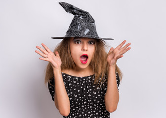 Happy halloween concept. Surprised Teen girl in witch black hat looking at camera in amazement on grey background. Cute child in witch halloween costume shouting, opening eyes and mouth with shock.