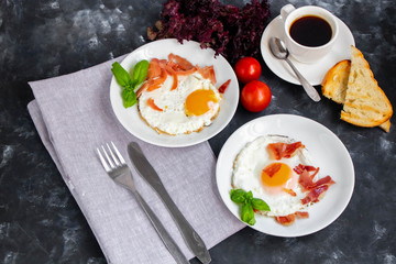 Breakfast. Fried eggs with bacon decorated with basil leaves. Nearby is a cup of coffee with toast. Chery tomatoes, gray napkin and cutlery. All on a dark background. 