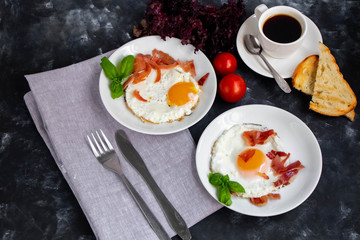 Breakfast. Fried eggs with bacon decorated with basil leaves. Nearby is a cup of coffee with toast. Chery tomatoes, gray napkin and cutlery. All on a dark background. 