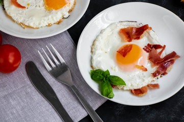 Breakfast. Fried eggs with bacon decorated with basil leaves. Nearby is a cup of coffee with toast. Chery tomatoes, gray napkin and cutlery. All on a dark background. 