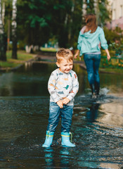 Child after a rain. Cute happy child boy is playing in the puddle. Young woman in jeans and boots in the background.