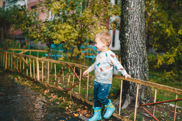 Little boy after the rain wearing rubber boots stand in big puddle near fence.