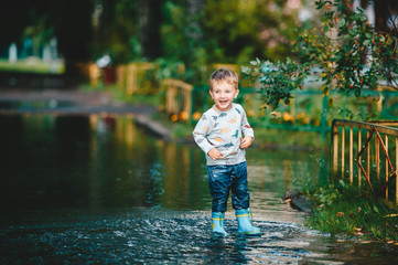 Naklejka premium Child after a rain. Cute happy child boy is playing in the puddle. Kid in black rubber boots.