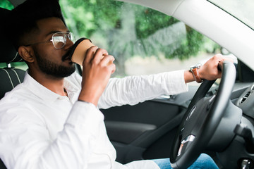 Transportation and vehicle concept. Young indian man drinking coffee while driving the car