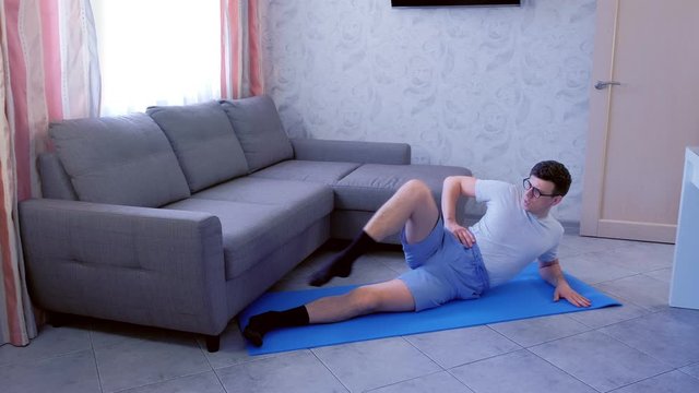 Weak nerd man in glasses is doing funny pilates exercises for legs laying on the floor at home. Sport humor concept.
