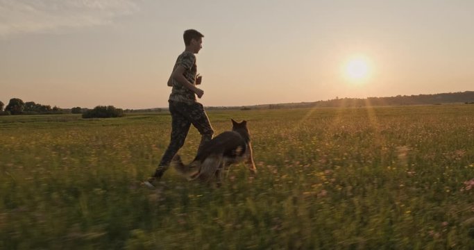 Teenager is running with a dog in nature while sunset. 15 year old boy runs with a dog breed German shepherd. Teen on the field with dog. Caucasian guy is playing with his pet in the meadow. 