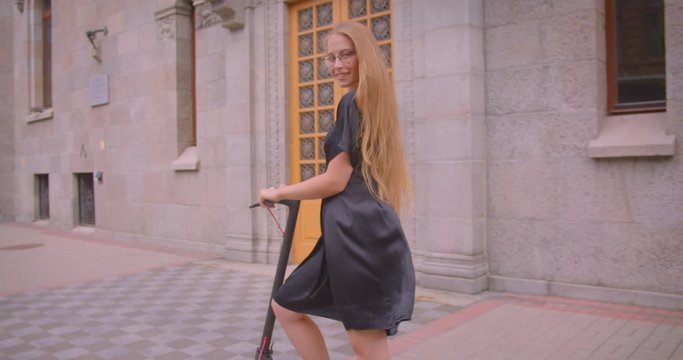 Closeup portrait of young attractive caucasian female with long hair in glasses and dress holding kick scooter looking at camera outdoors