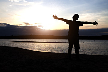 Fototapeta premium Silhouette of boy with arms spread out next to river sunset. Child on river sunset.