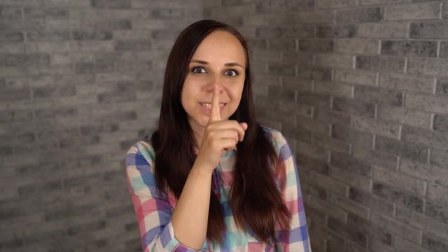 A beautiful emotional woman in a plaid shirt is doing silencing gesture with index finger at her lips on a gray brick background. The concept of silence.
