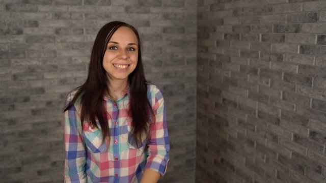 A beautiful emotional woman in a plaid shirt is doing silencing gesture with index finger at her lips on a gray brick background. The concept of silence.