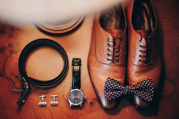 Stylish watch, expensive, shoes, bow tie, cufflinks and belt for groom on wooden table in hotel...